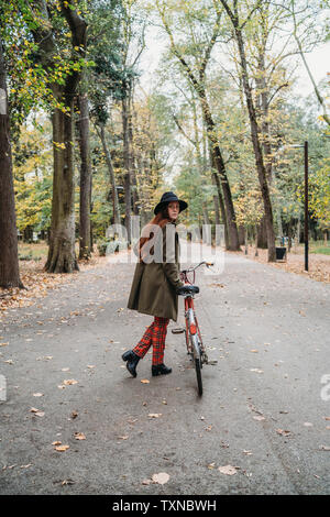 Junge Frau mit langen roten Haaren schaut über ihre Schulter, während Fahrrad schieben im Herbst Park, volle Länge, Florenz, Toskana, Italien Stockfoto