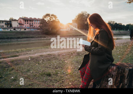 Junge Frau mit langen roten Haaren auf Baumstumpf lesen Buch über Riverside bei Sonnenuntergang, Florenz, Toskana, Italien sitzen Stockfoto