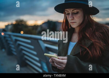 Junge Frau mit langen roten Haaren auf der Fußgängerbrücke auf Smartphone bei Dämmerung, Florenz, Toskana, Italien Stockfoto