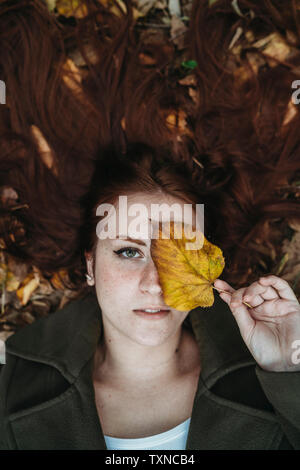 Junge Frau mit langen roten Haaren liegen unter Blätter im Herbst und das Auge mit Herbst Blatt, Overhead portrait