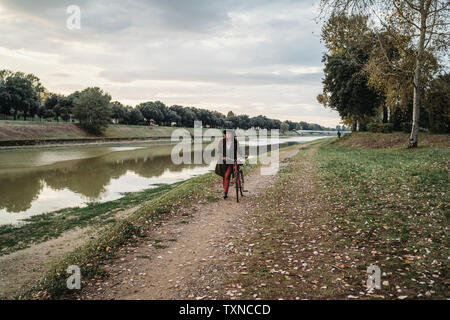 Junge Frau mit langen roten Haaren drücken Fahrrad auf Riverside, volle Länge, Florenz, Toskana, Italien Stockfoto