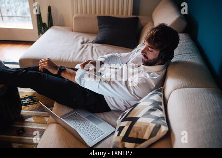 Mitte der erwachsene Mann liegend auf dem Sofa hören von Kopfhörern beim Ausrollen der Ärmel Stockfoto