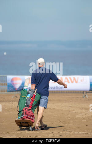 Mann in Sommershorts, offene Sandalen & Sportmütze, Strandwagen am Strand entlang ziehen, Weston-super-Mare, Großbritannien. Vater holt Strandausrüstung. Stockfoto