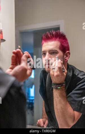 Diverse europäische männlich Mann mit pfennigabsatz rosa Haare Anwendung eyeliner Make-up im Spiegel Reflexion. Das Tragen von schwarzer Kleidung mit Punk Rocker alternative l Stockfoto