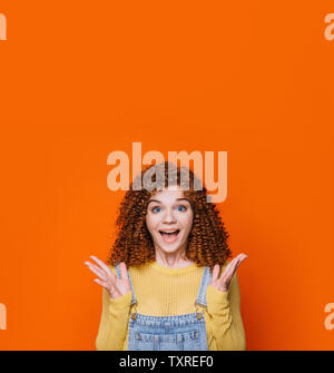 Curly rothaarige Frau, die schockiert Gesicht und hob die Hände auf orangem Hintergrund. Wow es ist erstaunlich Stockfoto