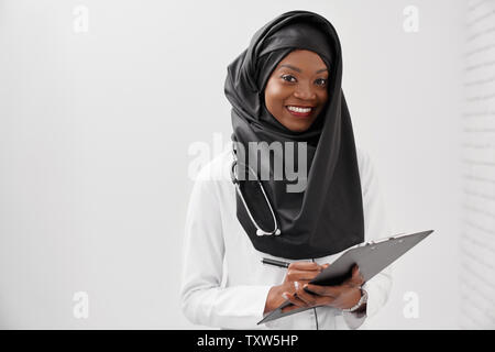 Schöne, positive weibliche Arzt mit Stethoskop Ordner und Stift, an der Kamera schaut. Fröhliche Frau hijab tragen in schwarz und weißen Mantel schreiben und lächelnd. Stockfoto