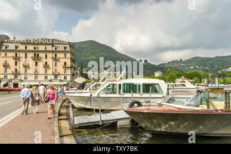 COMO, ITALIEN - JUNI 2019: Menschen zu Fuß am Seeufer mit Motor Boote neben in Como am Comer See. Im Hintergrund ist das Hotel Metropole Suisse Stockfoto