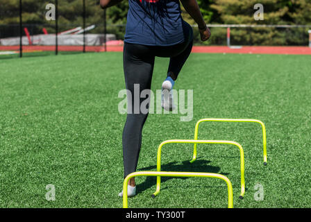 Ein High School Mädchen ist die Geschwindigkeit und Agilität Bohrer über Hürden ohne Schuhe auf, nur Socken auf einen grünen Rasenfeld mit schwarzen spandex. Stockfoto