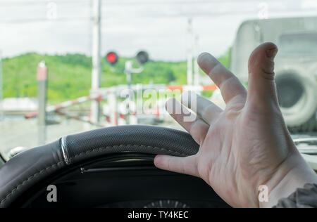 Die Hand des Mannes im Auto. Der Wagen hielt vor einer geschlossenen Schranke und eine rote Ampel vor dem Bahnübergang. Mann empört über die Stockfoto