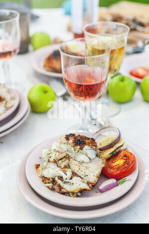 Gegrillte Zackenbarsch, Brille mit weißen und rosa Wein, Teller, Gemüse, Salat und Obst auf den Tisch. Sommerfest im Hinterhof. Vertikaler Dampfstoß Stockfoto