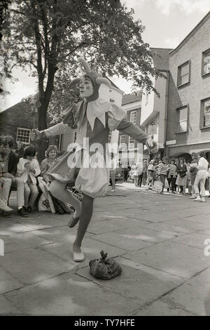 80er Jahre, eine moderne Hofnarr oder Entertainer Durchführung außerhalb in einer Straße, in ein Kostüm der ähneln, ihre historischen Pendants gekleidet. England, UK. Im Mittelalter, ein Spaßvogel war ein Schauspieler, der gemeinsamen Folk auf Messen und Märkten unterhalten, während der "hofnarr" Mitglied des königlichen Haushalts, deren Job war der König und seine Gäste in Bankette und Partys zu unterhalten wurde. Narren haben eine lange Geschichte und wurden in vielen von Shakespeares Dramen, einschließlich 'Twelfth Night', wo Feste der Narr ist beschrieben als "klug genug, der Narr" zu spielen. Stockfoto