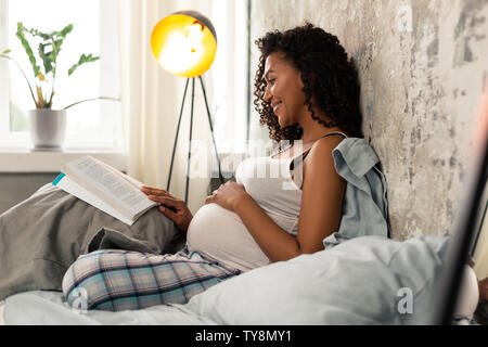 Fröhliche schwangere Frau mit einem Buch in ihrem Bett. Stockfoto