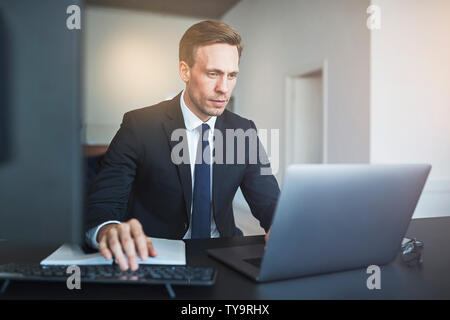 Konzentrierte Geschäftsmann online arbeiten mit einem Laptop sitzend an seinem Schreibtisch in einem hellen modernen Büro Stockfoto