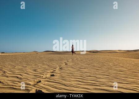 Eindrucksvolle junge selbstbewusste Frau zu Fuß auf den Weg in die Wüste Sand mit roten Kleid in der middlw der Dünen an heissen Sommertag mit klaren blauen Himmel Stockfoto