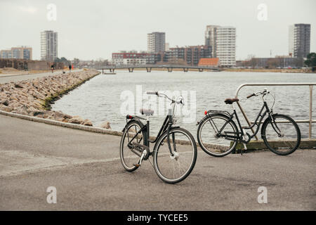 Zwei identische Retro schwarz Stadt Fahrräder am Bahndamm entlang der Straße geparkt, Ostsee, bahndamm mit Asphalt weg Hintergrund der Sdan und Stadt Copenhag Stockfoto