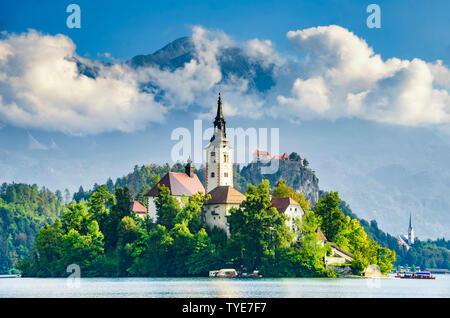 Tolle Aussicht mit wunderschönen Kirche und Schloss am See von Bled, Slowenien mit hoher Berge und Wolken im Hintergrund. Wallfahrtskirche der Assumpt Stockfoto