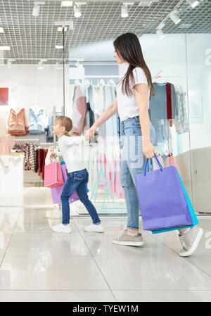 Seitenansicht der jungen attraktiven Mutter zusammen einkaufen mit kleiner Tochter in der Mall. Positive Mädchen die Hand der Frau und in Geschäften. Familie die Auswahl und Kauf neuer Kleidung und Spielzeug. Stockfoto