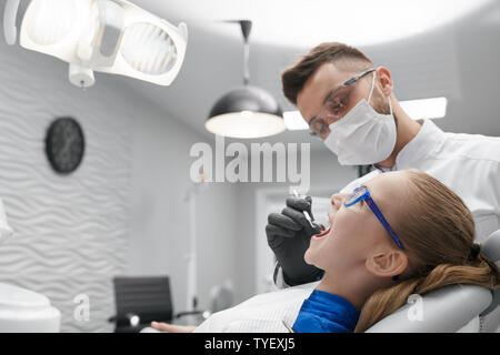 Seitenansicht der schweren männlichen Zahnarzt in Maske und Brille putzen die Zähne der kleinen Patienten in der Klinik. Hübsches Mädchen mit offenen Mund liegen auf zahnarztstuhl. Konzept der Behandlung und Gesundheit. Stockfoto