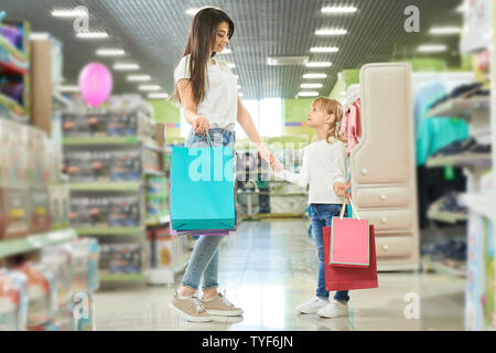 Junge Mutter zusammen einkaufen mit kleiner Tochter in der Mall. Glückliche Frau halten Beutel und die Hand des Mädchens beim Kauf neuer Spielsachen im Shop. Hübsche Brünette und Kind an jedem anderen zu schauen und zu reden. Stockfoto