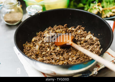 Der Küchenchef braten der in einer tiefen Pfanne hacken. Bereit, Füllung mit Zwiebeln in eine Schüssel auf dem Hintergrund von Gewürzen und Grüns. Das gebratene Fleisch Rühren mit Holzkasten Stockfoto