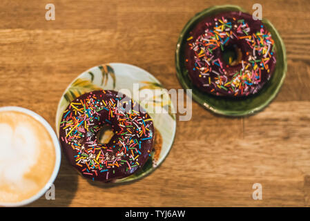 Schlechtes Frühstück. Kaffee mit einem gezeichneten Herz und Milch auf einen hölzernen Tisch in einem Coffee Shop. zwei Schokolade Donuts mit Streuung auf den Tisch neben der Co Stockfoto