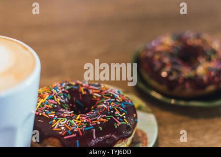 Schlechtes Frühstück. Kaffee mit einem gezeichneten Herz und Milch auf einen hölzernen Tisch in einem Coffee Shop. zwei Schokolade Donuts mit Streuung auf den Tisch neben der Co Stockfoto