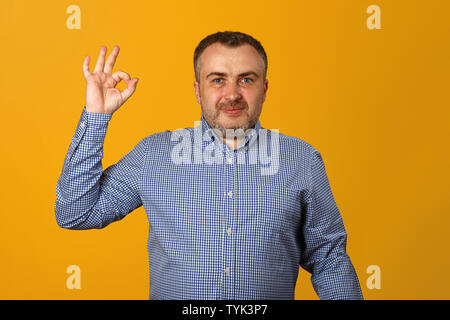 Ein junger Mann in einem T-Shirt zeigt das OK-Zeichen und sieht in die Kamera. Gelber Hintergrund. Close-up. Stockfoto