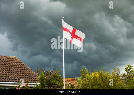 Englische Flagge von St. George's Cross auf einem Fahnenmast, neben einem Haus Dach, die sich gegen die dunklen Wolken einer stürmischen Himmel. England, UK. Stockfoto