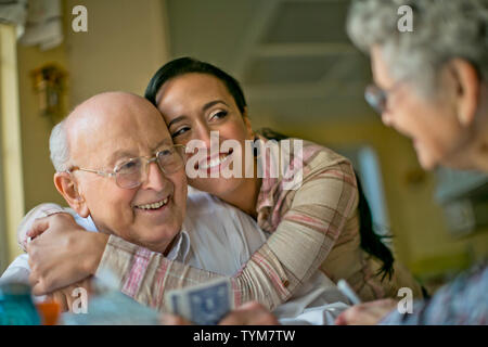 Lächelnde Frau mittleren Alters umfaßt ihr fröhlichen Großvater als ihre älteren Großeltern spielen Karten gemeinsam am Küchentisch. Stockfoto