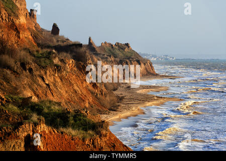 Stürmische See mit leuchtend roten und gelben Felsen an der Küste von Sonne beleuchtet Stockfoto