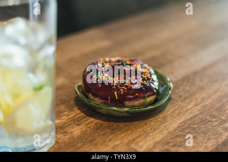 Kalte Limonade mit Eis und Zitronen in ein hohes Glas mit einem Einweg-röhrchen. Schokolade Donut mit Ankleideraum. Lecker und ungesunde Frühstück. Stockfoto