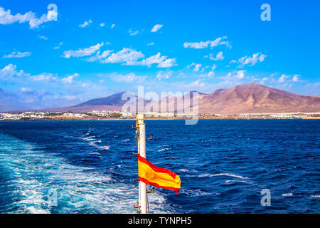 Spanische Flagge auf einem Kreuzfahrtschiff auf See. Im Hintergrund sind Lanzarote, Kanarische Inseln. Es gibt blaue Wasser des Ozeans. Stockfoto