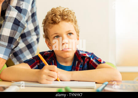 Während der Lektion. Ernsthafte blonde Junge, gelangweilt, während seine Aufgabe schreiben während der Lektion Stockfoto