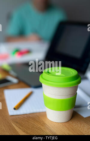 Ein Bambus wiederverwendbare Kaffeetasse sitzt auf einem hölzernen Tisch zusammen mit anderen Elementen wie Text Bücher und ein Laptop, der vorschlägt, es ist jemand der Studie. Stockfoto