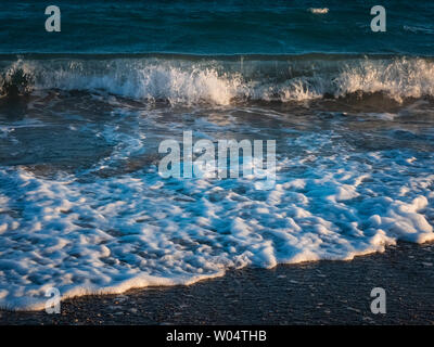 Spritzer schäumenden Wellen des Meeres Beat am Kiesstrand in den Strahlen der untergehenden Sonne. Stockfoto