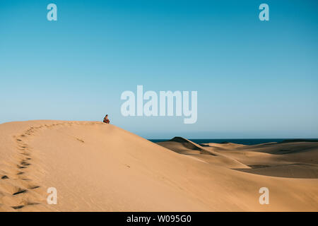 Junge Frau mit roten Kleid sitzen allein in der Wüste denken und auf Dune widerspiegelt als Metapher von Einsamkeit und Depression Stockfoto