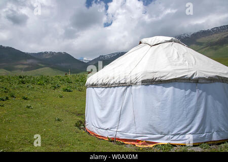 Jurte auf einer grünen Weide auf dem Hintergrund der Berge mit schneebedeckten Gipfeln und einem bewölkten Himmel. Das Reisen in Kirgistan. Stockfoto