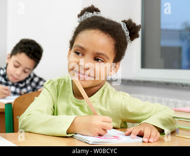 Hübsche Schülerin mit lockigem Haar Halten in der Hand pen Il, am Schreibtisch in der Nähe der Fenster sitzen, wegsehen, lächelnd, posieren. Kinder der Grundschule auf Lektion schreiben im Notebook. Stockfoto