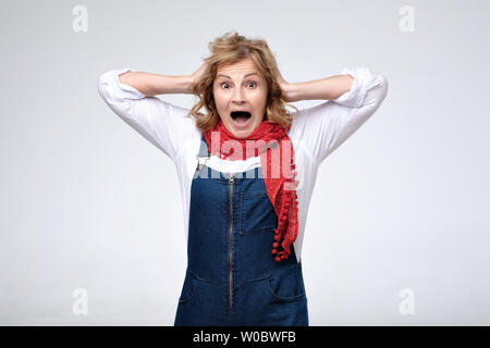 Extrem freundliche reife Frau tragen Jeans Overalls und roten Schal den Kopf berühren erstaunt, denn sie überrascht ist Stockfoto