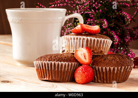Chocolate Muffins mit Erdbeeren, eine Tasse Kaffee und ein rosa Heather im Hintergrund auf einem Holztisch. Close-up. Stockfoto
