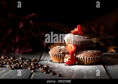 Chocolate Muffins mit Erdbeeren und Kaffee bestreut mit Puderzucker auf alten Dielen mit rosa Heather im Hintergrund. Stockfoto