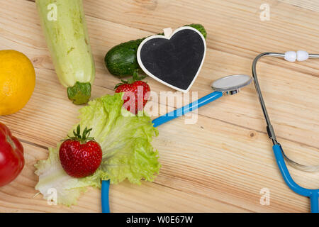 Gute Gesundheit und Diät Konzept - Tafel in Form von Herzen, Stethoskop und Gemüse, Früchte und Beeren auf hölzernen Tisch Stockfoto