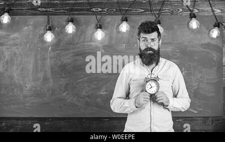 Lehrer in Brillen hält Wecker. Zeitplan und Regime Konzept. Bärtige hipster hält, schwarzen Brettes auf Hintergrund, kopieren. Mann mit Bart und Schnurrbart auf überrascht Gesicht im Klassenzimmer. Stockfoto