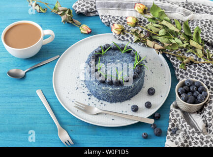 Tabelle mit blauen Blueberry Velvet Cake. Tasse Kaffee mit Milch, kleine Schüssel mit Blaubeeren, Schal und trockene Blumen, Rosen. Stockfoto