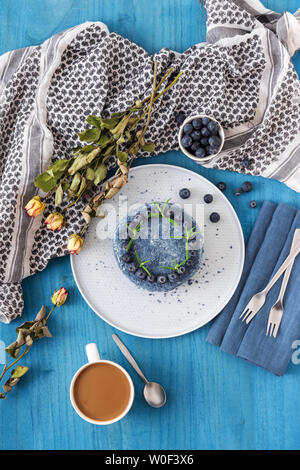Tabelle mit blauen Blueberry Velvet Cake. Tasse Kaffee mit Milch, kleine Schüssel mit Blaubeeren, Schal und trockene Blumen, Rosen. Stockfoto