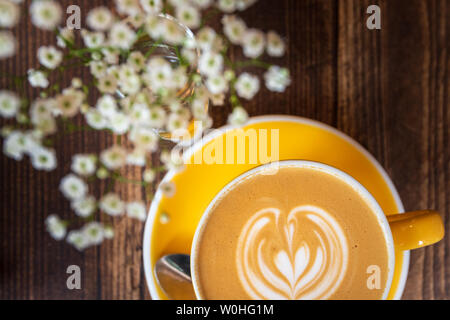 Latte Kaffe in hellem Gelb Tasse und Untertasse neben einigen Blumen auf einem Holztisch Stockfoto