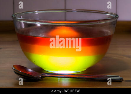 Nahaufnahme von einem großen Glas Schale mit drei Schicht Gelee / jello Nachtisch, bestehend aus Limone, Orange und Erdbeer, mit Löffel auf einem mit Pinien Küchenarbeitsplatte. Stockfoto