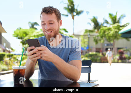Mann auf Cafe mit Smart Phone app Text Messaging sms trinken Eiskaffee im Sommer. Hübscher junger legerer Mann mit Smartphone lächelt glücklich Sitzen im Freien. Urban Mann in seinen 20ern. Stockfoto