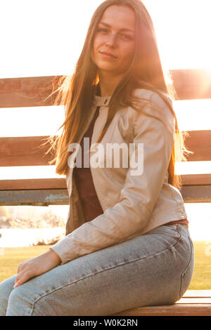 Schöne Mädchen mit langem Haar sitzt gegenüber dem Sonnenlicht in beige Lederjacke und Jeans Stockfoto