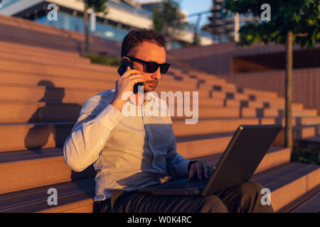 Seitenansicht Portrait von Young Business Executive Aufruf auf Smartphone und mit Laptop im Freien. Stockfoto